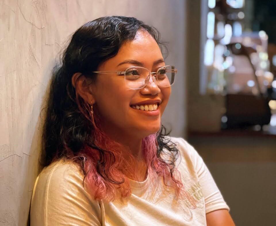 Whatsapp image 20260610 at 17 12 21 catrawarta Smiling woman with glasses sits against a textured wall in a warm indoor setting